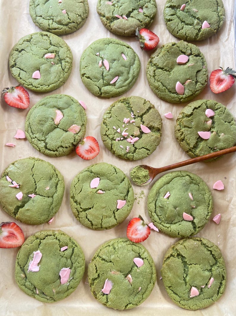 May include: A collection of round, green matcha cookies featuring pink chocolate chips and fresh strawberry slices. A small wooden spoon containing a mound of green matcha powder is positioned among the cookies on a sheet of parchment paper.