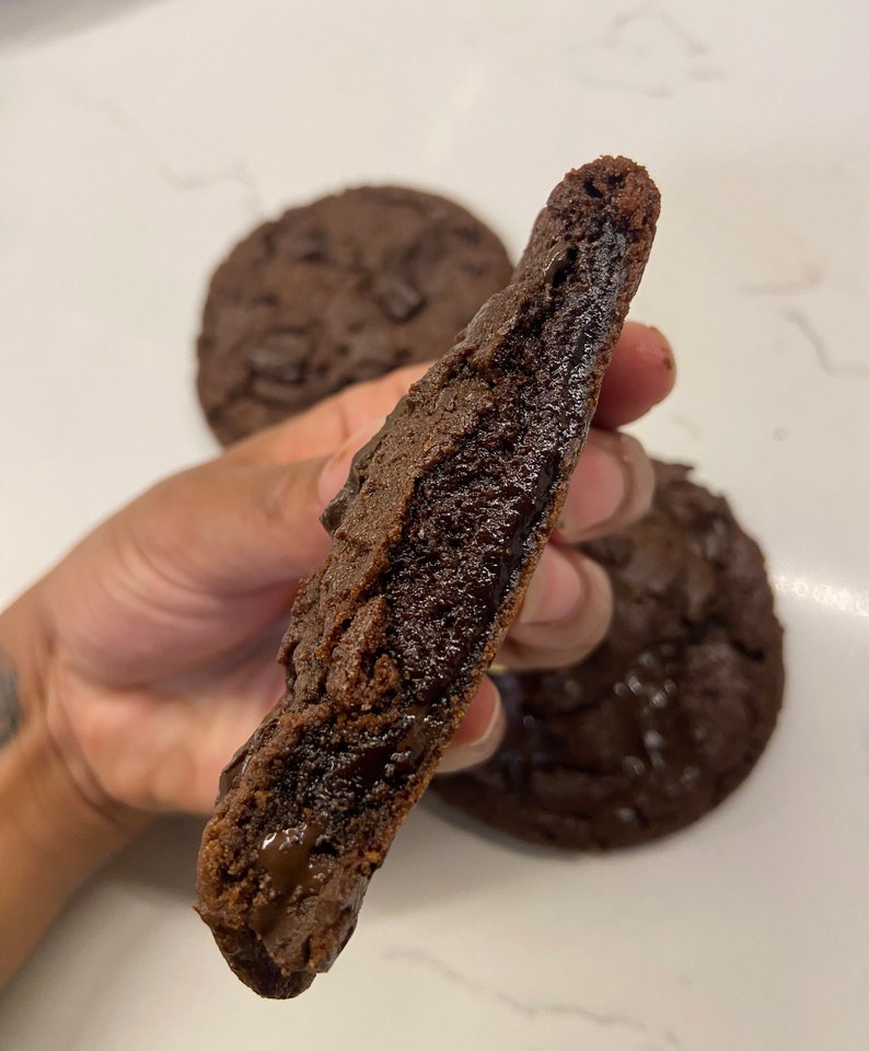 May include: A dark chocolate cookie broken in half to reveal a soft, molten, and gooey center. The cookie has a deep brown color with visible chunks of melted chocolate throughout the dough. Two additional whole chocolate cookies are visible.