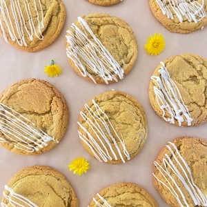 May include: A collection of eight round, golden-brown baked cookies arranged in a grid pattern. Each cookie features a textured surface and is decorated with a drizzle of white icing. Three small, bright yellow dandelion flowers are interspersed.