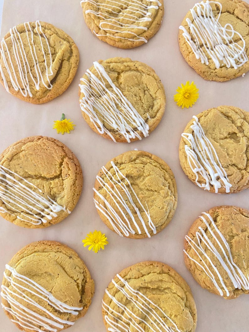 May include: A collection of eight round, golden-brown baked cookies arranged in a grid pattern. Each cookie features a textured surface and is decorated with a drizzle of white icing. Three small, bright yellow dandelion flowers are interspersed.
