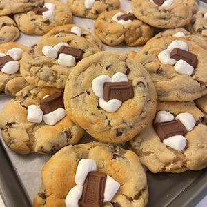 May include: A batch of golden-brown baked cookies arranged on a metal tray. Each cookie features a soft, round dough base with visible chocolate chips, topped with white marshmallows and a rectangular piece of Hershey's milk chocolate.