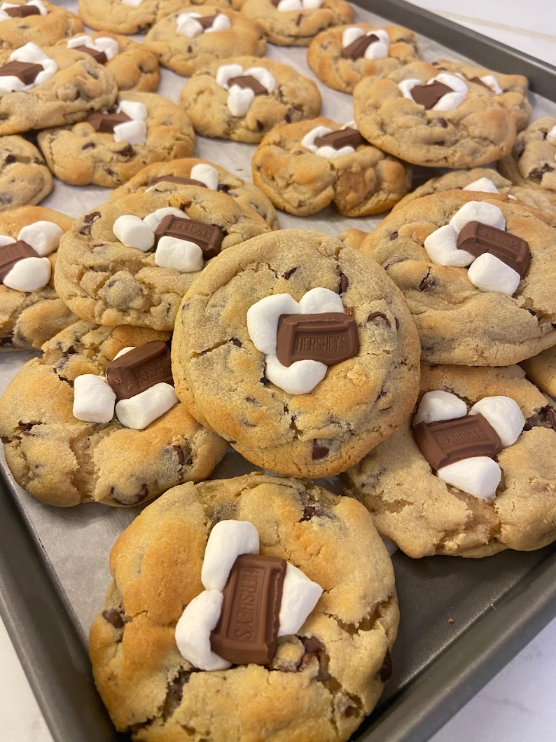 May include: A batch of golden-brown baked cookies arranged on a metal tray. Each cookie features a soft, round dough base with visible chocolate chips, topped with white marshmallows and a rectangular piece of Hershey's milk chocolate.