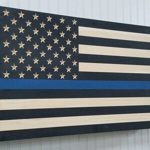 May include: A wooden American flag with a blue stripe across the middle, representing the thin blue line. The flag is painted in black and white with gold stars.