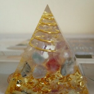 Op de afbeelding: Een heldere piramide met een gouden spiraal erin. De piramide is gevuld met kleine, kleurrijke kristallen en gouden vlokken.