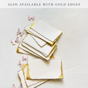 May include: A stack of white cards with gold edges. The cards are arranged in a fan shape and have a rough, textured surface. The cards are on a white background with small pink butterflies scattered around them.