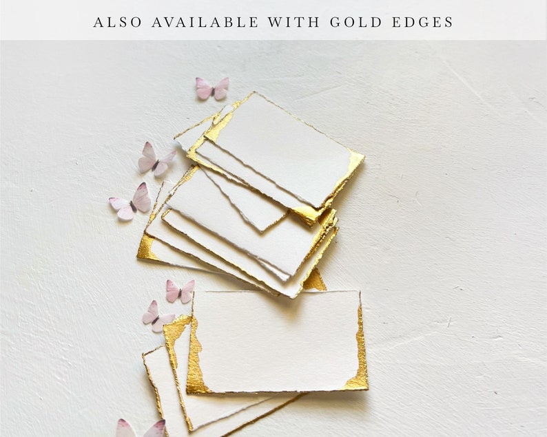 May include: A stack of white cards with gold edges. The cards are arranged in a fan shape and have a rough, textured surface. The cards are on a white background with small pink butterflies scattered around them.
