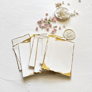 May include: A stack of white cards with gold edges, arranged on a white surface. The cards are blank and have a rough, textured surface. There are small pink flowers and a white wax seal with a leaf design scattered around the cards.