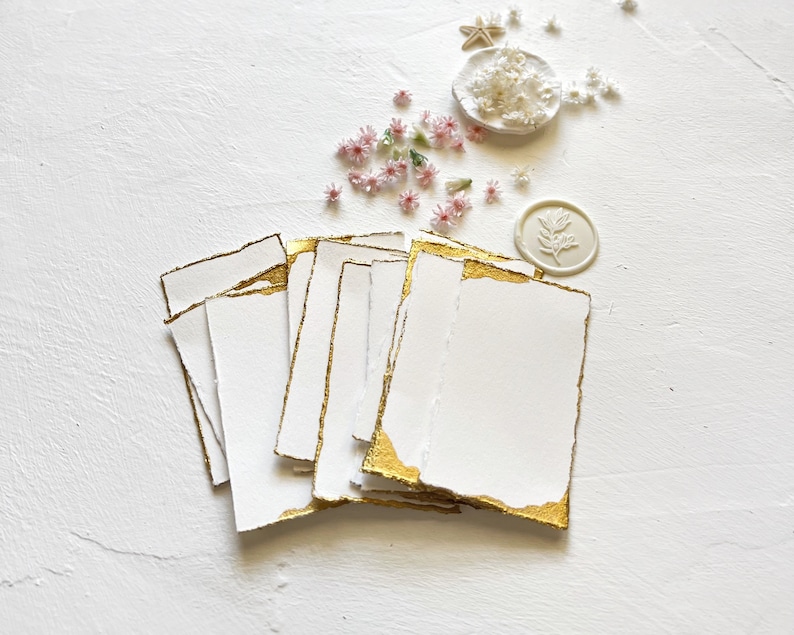 May include: A stack of white cards with gold edges, arranged on a white surface. The cards are blank and have a rough, textured surface. There are small pink flowers and a white wax seal with a leaf design scattered around the cards.