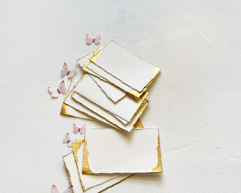 May include: A stack of white cards with gold edges, arranged in a fan shape. The cards are blank and have a rough, textured surface. Pink paper butterflies are scattered around the cards.