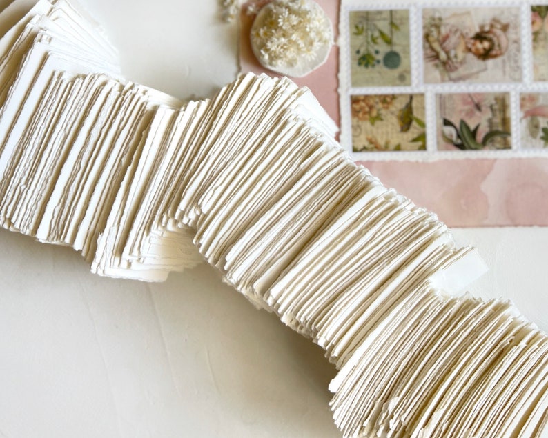 May include: A stack of white, handmade paper sheets with rough edges. The paper is arranged in a wave-like pattern.