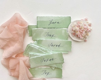 Op de afbeelding: Groene aquarel-kaartjes met namen geschreven in zwarte kalligrafie: Zara, Tony, Serah, Susan, Joy. Een klein wit bord met roze bloemen bevindt zich in de rechterbovenhoek.