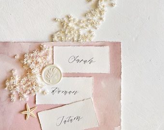 May include: Three white cards with calligraphy names on a pink background. The names are Sarah, Roman, and Tatum. There are white flowers and a white wax seal with a leaf design on the left side of the image.