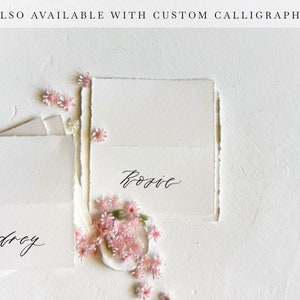 May include: Two white cards with black calligraphy. The top card says "Rosie" and the bottom card says "Audrey". Pink flowers are scattered around the cards.