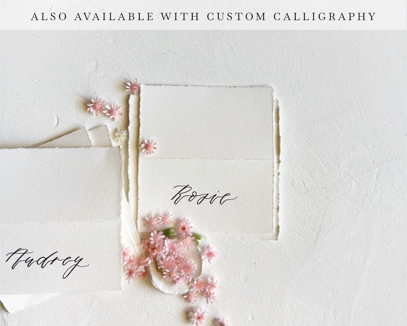 May include: Two white cards with black calligraphy. The top card says "Rosie" and the bottom card says "Audrey". Pink flowers are scattered around the cards.