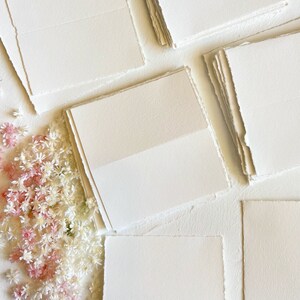 May include: A stack of white, textured paper cards with deckled edges. The cards are arranged in a fan shape and are surrounded by small white and pink flowers.