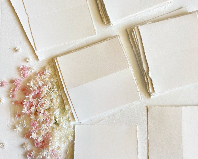 May include: A stack of white, textured paper cards with deckled edges. The cards are arranged in a fan shape and are surrounded by small white and pink flowers.