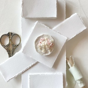 Op de afbeelding: Een wit, getextureerd papier met een delicate roze en witte bloemenarrangement in een ronde, witte lijst. Het papier wordt omringd door andere witte, getextureerde papieren en een antieke schaar.