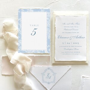 Pode incluir: Um convite de casamento azul e branco com um design floral. O convite inclui os nomes Eleanor e Arthur, a data 5.24.2025 e a hora 16h00. O convite tamb&eacute;m inclui a localiza&ccedil;&atilde;o, "Oliveta's Avenue, La Jolla, CA 92037".