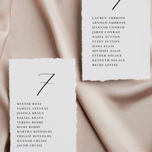 May include: Two white cards with black text listing names for a wedding reception seating chart. The cards are numbered "7" and are on a light pink satin background.