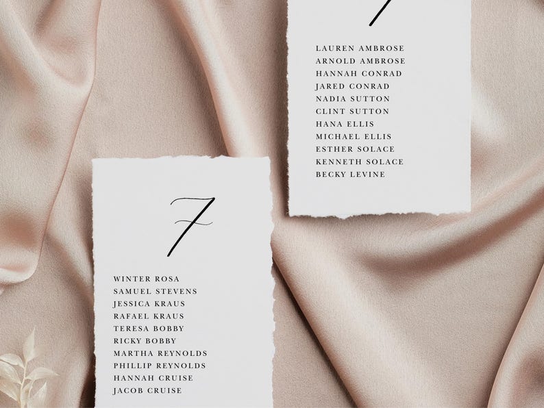 May include: Two white cards with black text listing names for a wedding reception seating chart. The cards are numbered "7" and are on a light pink satin background.