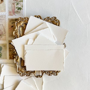 May include: A stack of white, textured, handmade paper cards with deckled edges, displayed in a gold ornate frame.