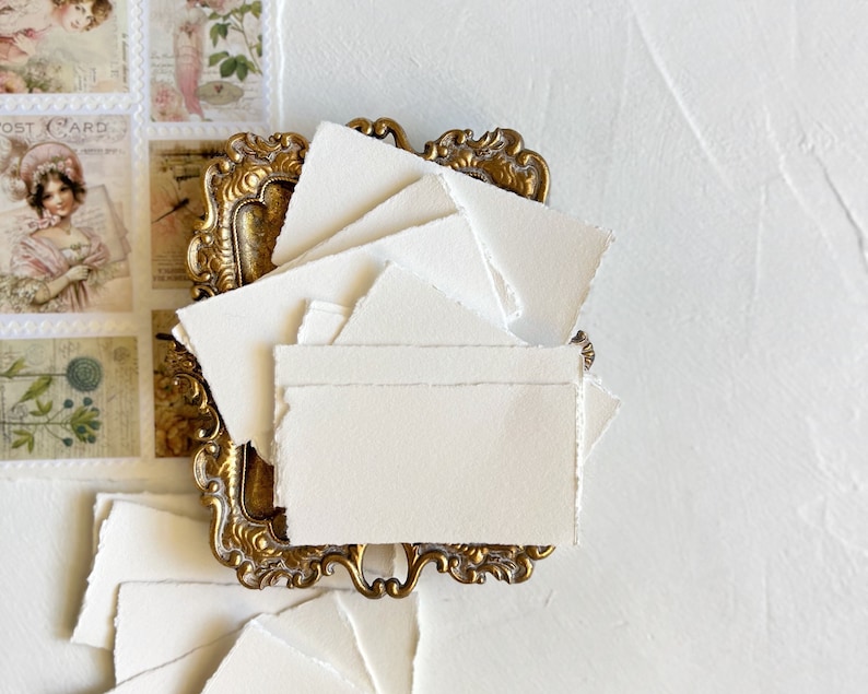 May include: A stack of white, textured, handmade paper cards with deckled edges, displayed in a gold ornate frame.