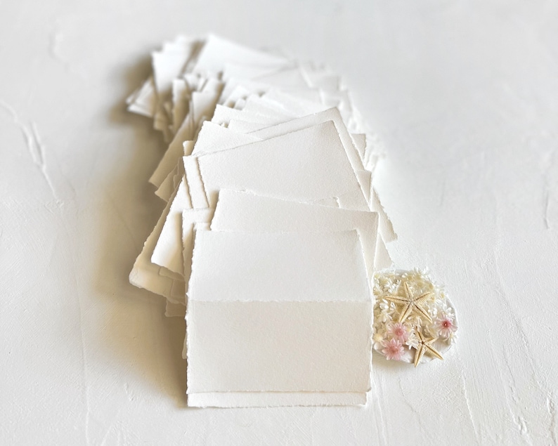 May include: A stack of blank, white, textured paper cards with deckled edges. The cards are arranged in a fan shape with the top card folded in half. A small cluster of white starfish and pink flowers is in the lower right corner of the image.