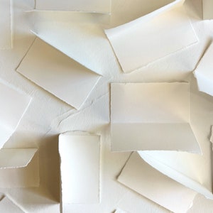 May include: A collection of white paper cards with deckled edges, arranged in a layered pattern on a white textured surface.