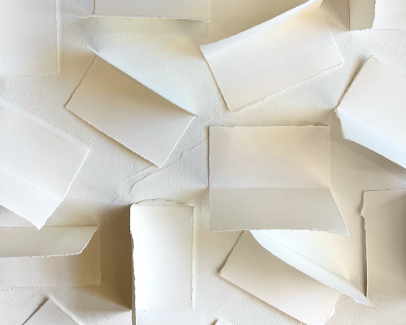 May include: A collection of white paper cards with deckled edges, arranged in a layered pattern on a white textured surface.