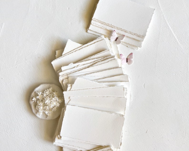 May include: A stack of white, handmade paper with deckled edges. The paper is arranged in a fan shape with two small pink butterflies on top. A small, round, white object with white flowers is in the lower left corner.