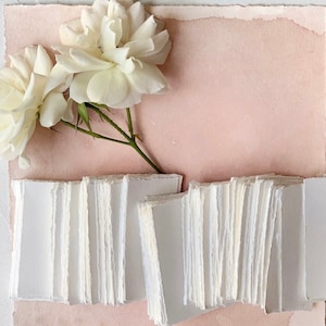 May include: A stack of white, handmade paper cards with deckled edges, arranged on a light pink background with white roses.