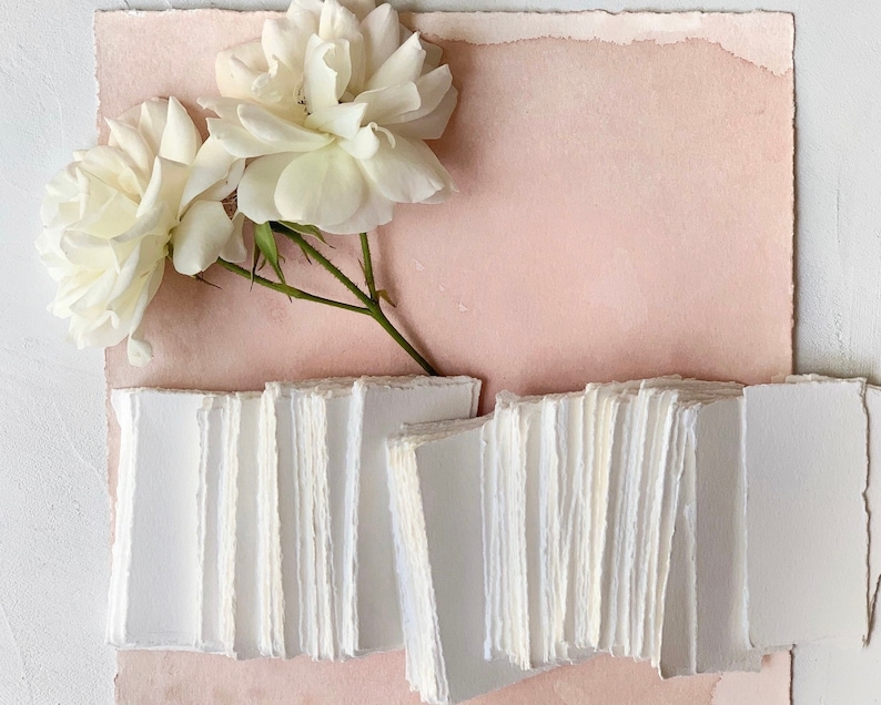 May include: A stack of white, handmade paper cards with deckled edges, arranged on a light pink background with white roses.