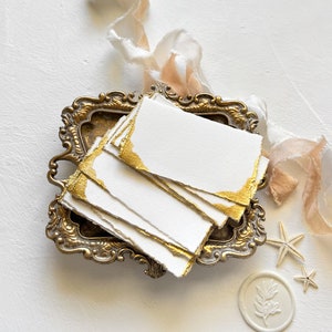 May include: A stack of white paper cards with gold edges, displayed in a vintage brass tray. The cards are surrounded by delicate ribbons and seashells.