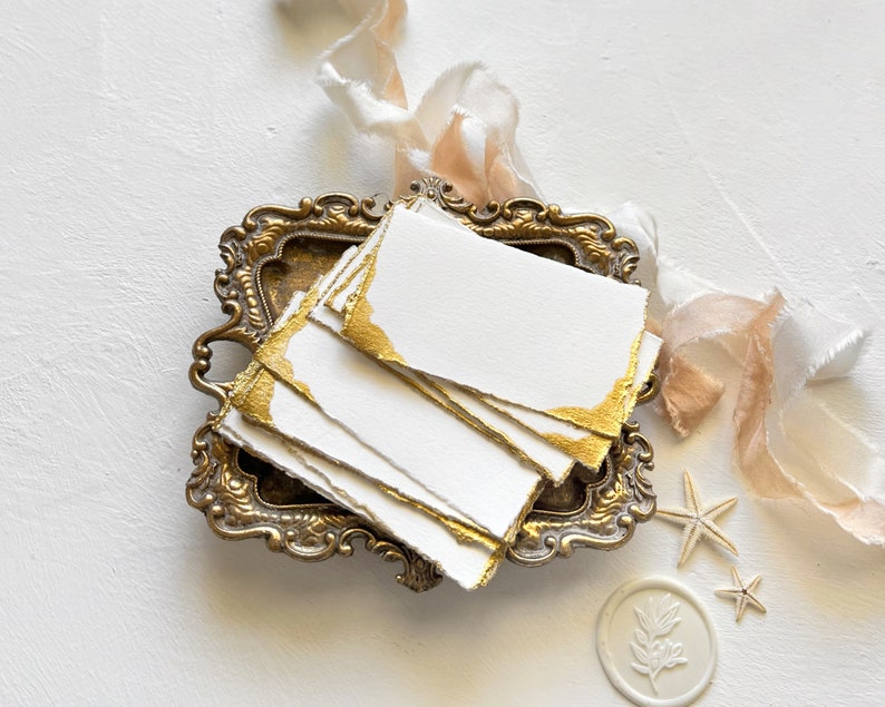 May include: A stack of white paper cards with gold edges, displayed in a vintage brass tray. The cards are surrounded by delicate ribbons and seashells.