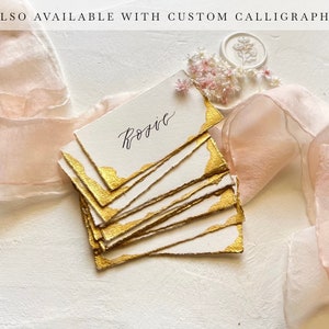 May include: A stack of white place cards with gold edges and the name "Rosie" written in calligraphy. The cards are on a white surface with a pink ribbon and small white flowers.