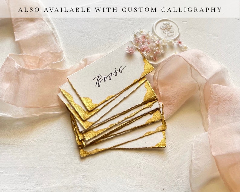 May include: A stack of white place cards with gold edges and the name "Rosie" written in calligraphy. The cards are on a white surface with a pink ribbon and small white flowers.