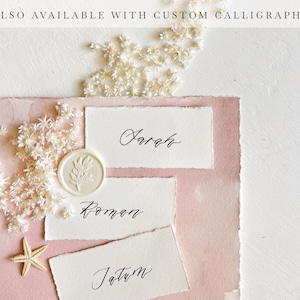 May include: Three white place cards with calligraphy names "Sarah", "Roman", and "Jatum" on a pink background with white flowers and a white wax seal.