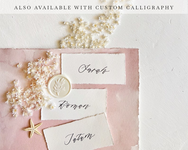 May include: Three white place cards with calligraphy names "Sarah", "Roman", and "Jatum" on a pink background with white flowers and a white wax seal.