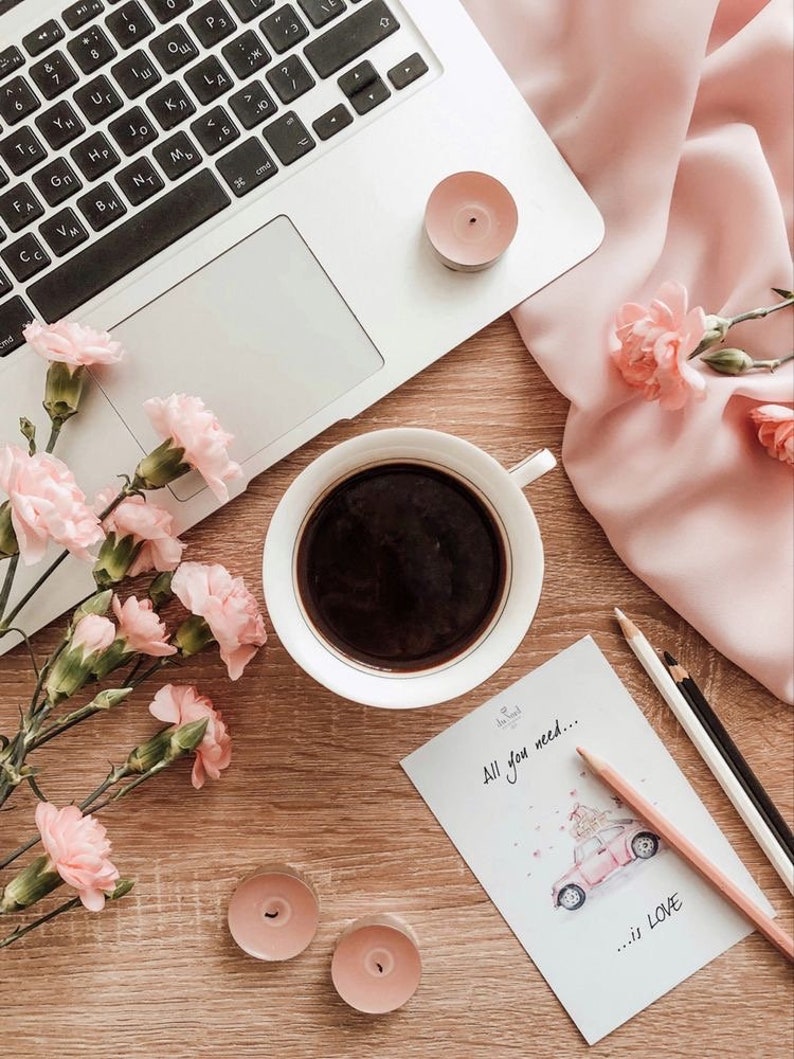 Op de afbeelding: Een platte lay-out van een houten tafel met een laptop, een kopje koffie, roze bloemen, kaarsen en een kaart met de tekst "Alles wat je nodig hebt... is LIEFDE".