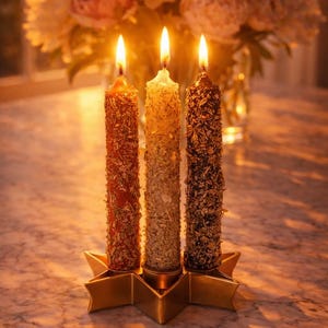 May include: Three lit, pillar candles in shades of red, gold, and brown, adorned with decorative elements, sit in a gold star-shaped holder. A bouquet of flowers is in the background. The candles are on a marble surface.