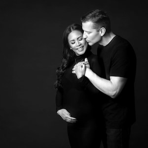 May include: A black and white photo of a couple embracing. The woman is pregnant and the man is kissing her on the cheek. They are both smiling.