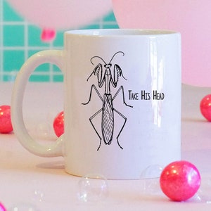 May include: White ceramic mug with a black and white illustration of a praying mantis and the text "Take His Head". The mug is surrounded by pink and white balloons and bubble wrap.