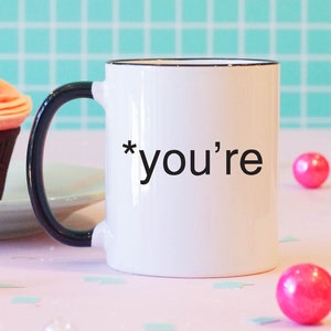 May include: White coffee mug with a black handle and black rim. The mug has the text "*you're" printed on it. A pink cupcake with a lit candle is on a plate in the background.