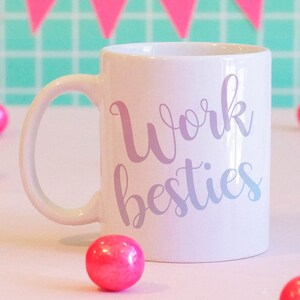 Pu&ograve; includere: Una tazza in ceramica bianca con un design viola e bianco che dice "Work besties". La tazza &egrave; circondata da gomme da masticare rosa su una superficie rosa.