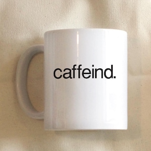 May include: White ceramic mug with a black inscription that reads "caffeind." The mug is sitting on a beige fabric surface next to a slice of grapefruit.