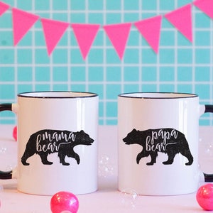 May include: Two white ceramic mugs with black handles and rims. Each mug features a black bear silhouette with the words "mama bear" and "papa bear" in cursive. Pink bubble-shaped decorations and a pink triangle banner are in the background.