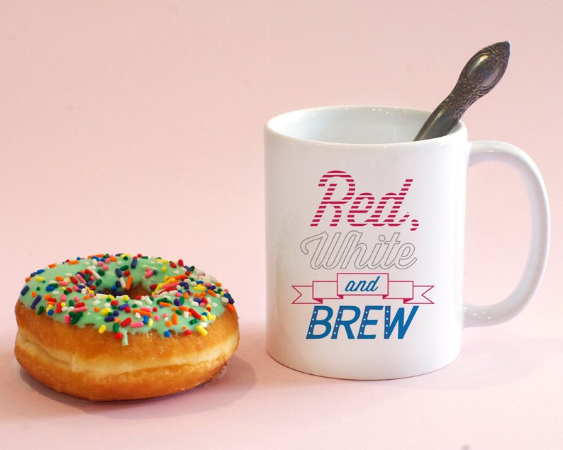 May include: White ceramic mug with red, white and blue text that reads "Red, White and BREW". A green glazed donut with colourful sprinkles is in the foreground.