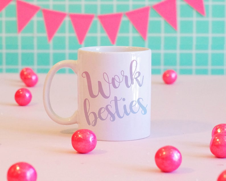Pu&ograve; includere: Una tazza in ceramica bianca con un design viola e bianco che dice "Work besties". La tazza &egrave; circondata da gomme da masticare rosa su una superficie rosa.