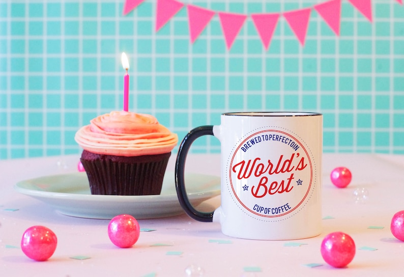May include: White coffee mug with a black handle and a red and blue circle design that says "Brewed to Perfection World's Best Cup of Coffee". The mug is on a white table with a pink cupcake and pink gumballs.