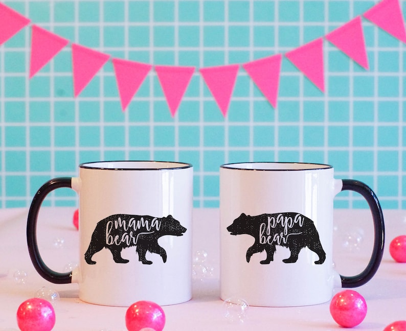 May include: Two white ceramic mugs with black handles and rims. Each mug features a black bear silhouette with the words "mama bear" and "papa bear" in cursive. Pink bubble-shaped decorations and a pink triangle banner are in the background.
